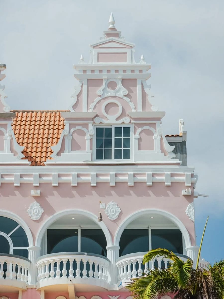 A pink colonial building in Oranjestad with white ornate gables, round windows and a palm tree