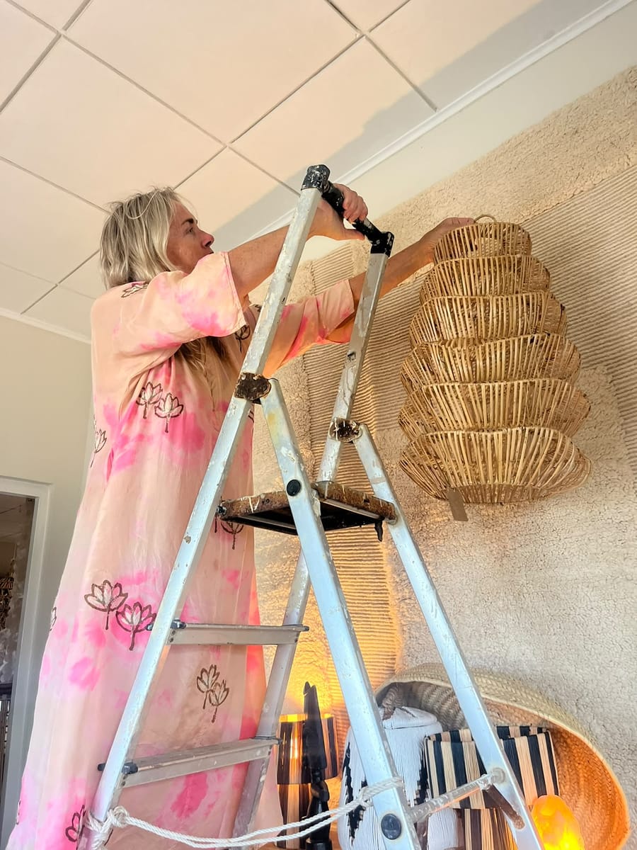 Woman in a pink tie-dye dress on a ladder hanging a rattan lamp on a textured wall
