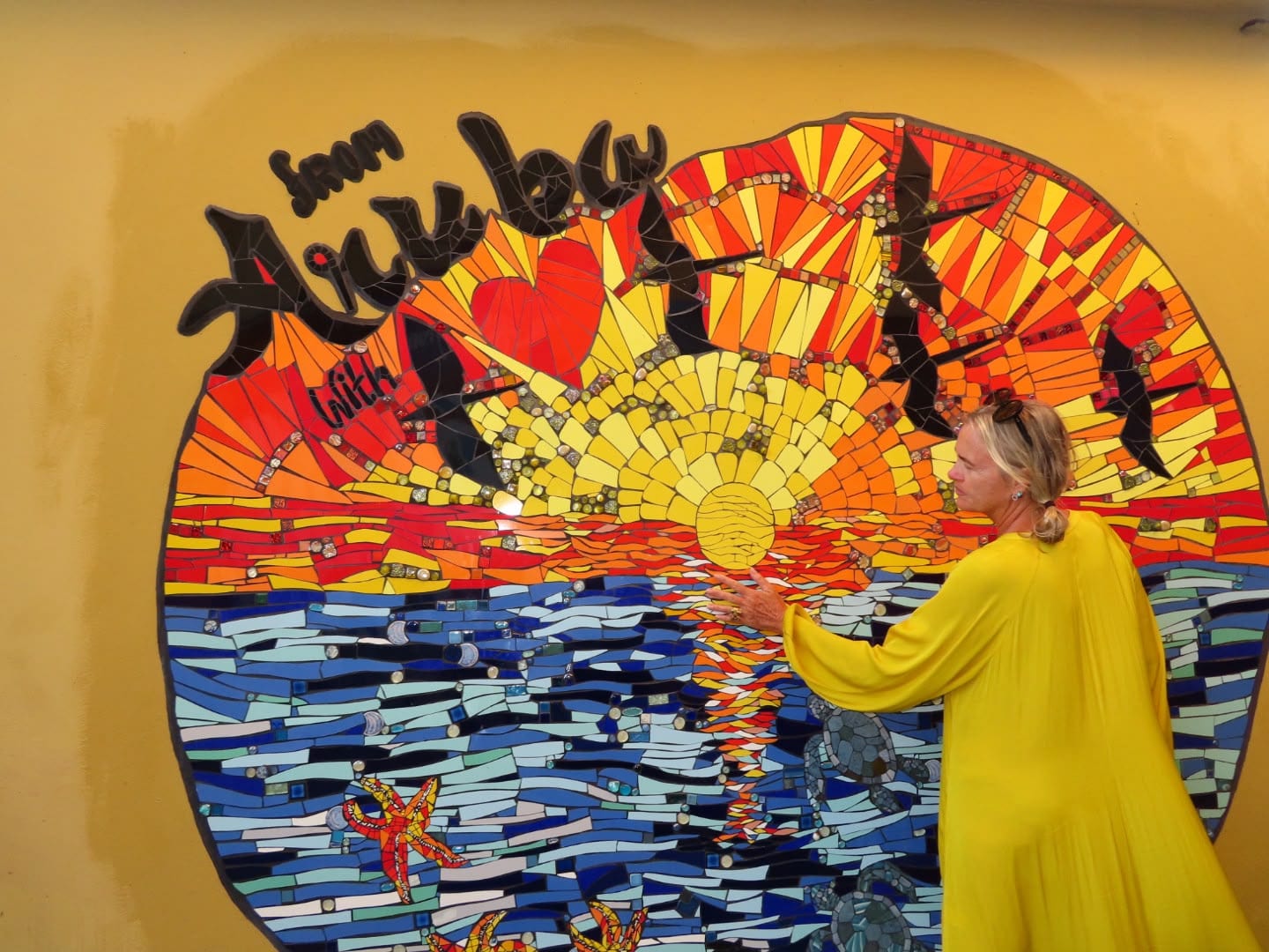 Woman in a yellow dress touching a colourful mosaic of a sunset over the sea